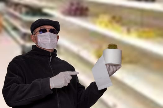 Coronavirus panic concept. Man with surgical mask, hood, glasses and medical gloves shows a roll of toilet paper with an abstract model of a virus covid-19 on it against blurred super market background.