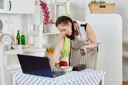 Woman in an apron with notebook and phone cooks in the kitchen. Isolation period, quarantine, social distancing. Remote education or remote work