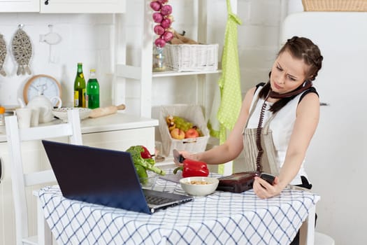 Woman in an apron with notebook and phone cooks in the kitchen. Isolation period, quarantine, social distancing. Remote education or remote work