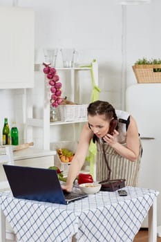 Woman in an apron with notebook and phone cooks in the kitchen. Isolation period, quarantine, social distancing. Remote education or remote work