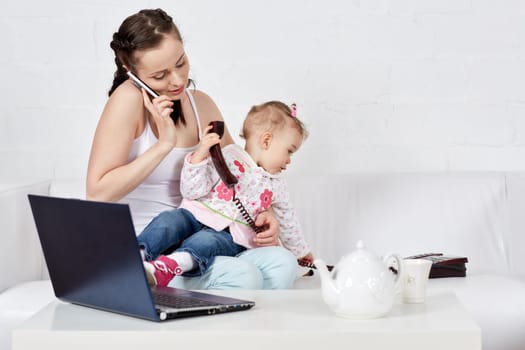 Small child and busy young woman with notebook and phone sit on the sofa in the room. Mother and baby. Isolation period, quarantine, social distancing. Remote work
