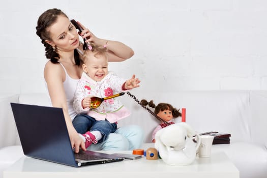 Small child and busy young woman with notebook and phone sit on the sofa in the room. Mother and baby. Isolation period, quarantine, social distancing. Remote work