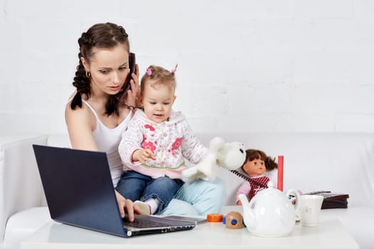 Small child and busy young woman with notebook and phone sit on the sofa in the room. Mother and baby. Isolation period, quarantine, social distancing. Remote work