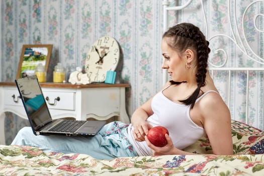 Young woman with laptop sits  on the house bed in the room. Isolation period, quarantine, social distancing. Remote education or remote work.