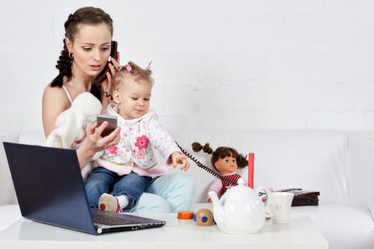 Small child and busy young woman with notebook and phone sit on the sofa in the room. Mother and baby. Isolation period, quarantine, social distancing. Remote work
