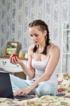 Young woman with laptop sits  on the house bed in the room. Isolation period, quarantine, social distancing. Remote education or remote work.
