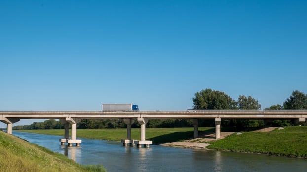 Bridge view and truck on the bridge. Pictured is a blue sky and a river, and a truck crossing the bridge over the river.