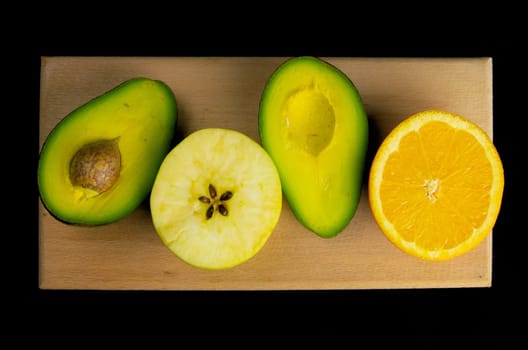 The concept of healthy diet and vegetarianism. Sliced avocado, apple and orange on a wooden board isolated on black background. Flat lay food.