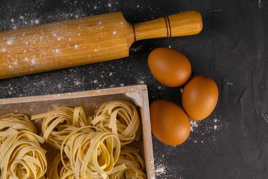 Still life of preparing pasta on rustic wooden background. Raw tagliatelle in a wooden box with rolling pin for dough and eggs. Top views.
