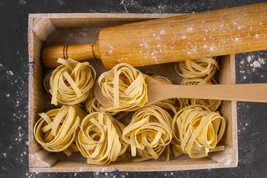 Still life of preparing pasta on rustic wooden background. Raw tagliatelle in a wooden box with rolling pin for dough and wooden spoon. Top views close-up