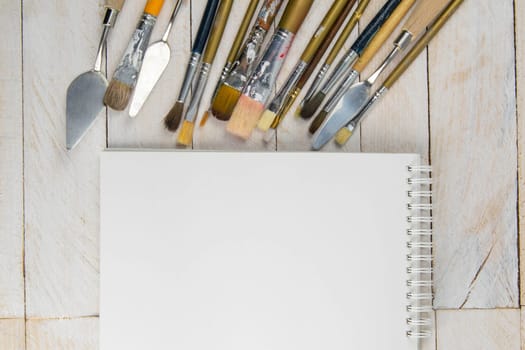 Set of artist accessories collection. Canvas, tube of oil paint, art brushes, palette knife and white open notebook lying on the wood table. Artist workshop background.