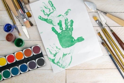 Set of artist accessories collection. Canvas, tube of oil paint, art brushes, palette knife and a letter with a print of children's hands in green paint lying on the wood table. Artist workshop background.