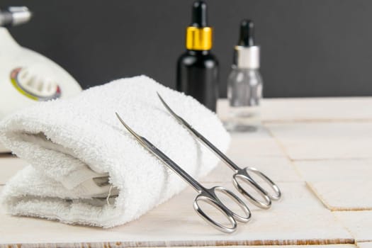 A set of cosmetic tools for manicure and pedicure on a blue background. Fraser, manicure scissors propped up to white towel, in the background are cuticoil oil and hand oil stand on a white wooden background.