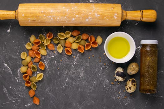 Pasta Conchiglie Rigate, mix of colorful uncooked shells with rocking for dough, olive oil, spices and quail eggs. Textured Italian food background concept. Top view flat lay close up, clear space.