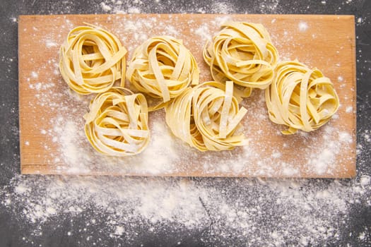 Variety of italian homemade raw uncooked pasta spaghetti and tagliatelle with flour on wooden board over black texture background. Flat lay.