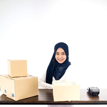E-Commerce asian muslim women merchant work from home on wooden table with postal parcel cardboard boxes for shipping orders. Studio shot on white background for Selling online and small business SME.