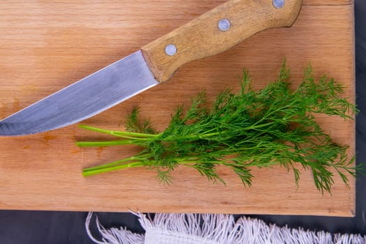 Fresh cut green dill and metal knife on wooden chopping board on white towel and black grunge kitchen table background. Cutting greens for adding to soup or salad. Healthy food.