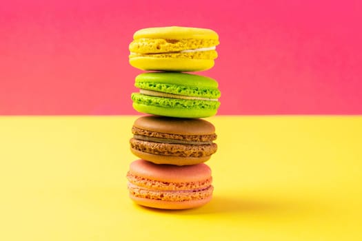 Multi-colored macaroons stand in a pile on a yellow table near a pink background. Close-up