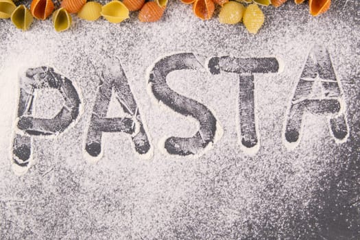 The words PASTA drawn a finger on flour with a kind of pasta, conchiglie. Italian pasta, flat lay close-up.