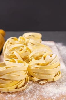 Variety of italian homemade raw uncooked pasta spaghetti and tagliatelle with flour on wooden board over black texture background. Front views.