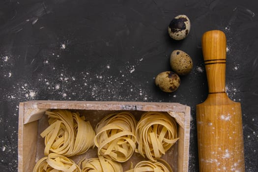 Still life of preparing pasta on rustic wooden background. Raw tagliatelle in a wooden box with rolling pin for dough and quail eggs. Top views.