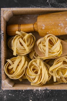 Still life of preparing pasta on rustic wooden background. Raw tagliatelle in a wooden box with rolling pin for dough. Top views close up