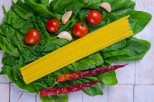 Raw spaghetti with tomatoes and spicy red pepper stand on spinach leaves on a white wooden table. Top views, close-up