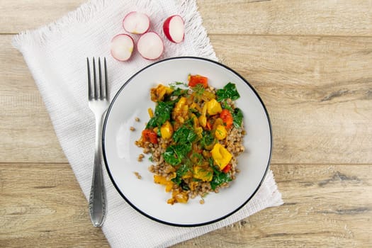 Delicious homemade vegetarian buckwheat with tomatoes, carrots, zucchini, yellow peppers and fresh basil on a light rustic wooden kitchen table. Top views, close-up