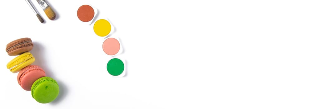 Makarons on a white background are next to the colors of macaroons and brushes for drawing. Top views with clear space.