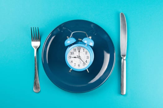 Alarm clock with fork and knife on the blue plate on blue table. Time to eat, Breakfast, Lunch Time and Dinner concept. Top views