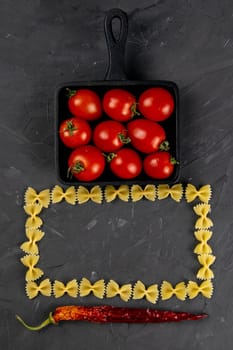 Raw pasta farfalle, tomatoes in frying pan and hot paper on dark background. Top views, close-up.