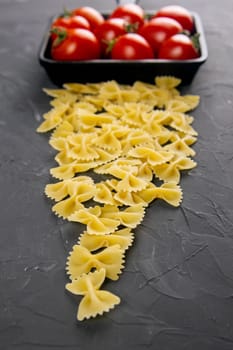 Raw pasta farfalle, tomatoes and frying pan on dark background. Top views, close-up.