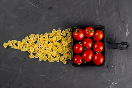 Raw pasta farfalle, tomatoes and frying pan on dark background. Top views, close-up.