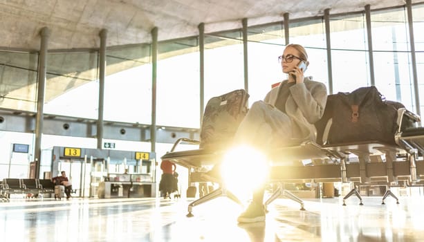 Casual blond young woman talking on cell phone while waiting to board a plane at airport departure gates.