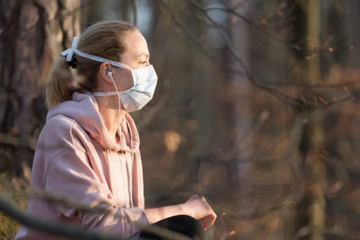 Portrait of caucasian sporty woman wearing medical protection face mask while relaxing in nature and listening to music. Corona virus, or Covid-19, is spreading all over the world.