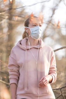 Portrait of caucasian sporty woman wearing medical protection face mask while walking in park, relaxing and listening to music. Corona virus, or Covid-19, is spreading all over the world.