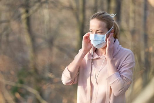 Portrait of caucasian sporty woman wearing medical protection face mask while walking in park, relaxing and listening to music. Corona virus, or Covid-19, is spreading all over the world.