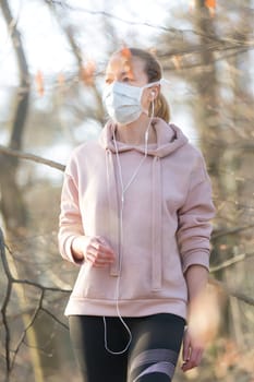 Portrait of caucasian sporty woman wearing medical protection face mask while walking in park, relaxing and listening to music. Corona virus, or Covid-19, is spreading all over the world.