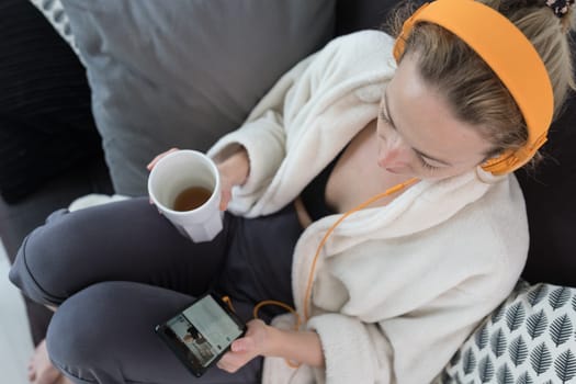 Woman at home relaxing on sofa couch drinking tea from white cup, listening to relaxing music while reading emails and using social media on mobile phone device. Stay at home. Social distancing.