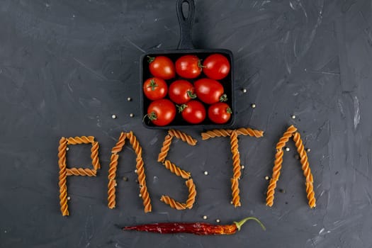 Italian fusilli - word pasta. Raw Italian pasta fusilli with cherry tomatoes in frying pan and hot paper on dark background from above. Flat lay.
