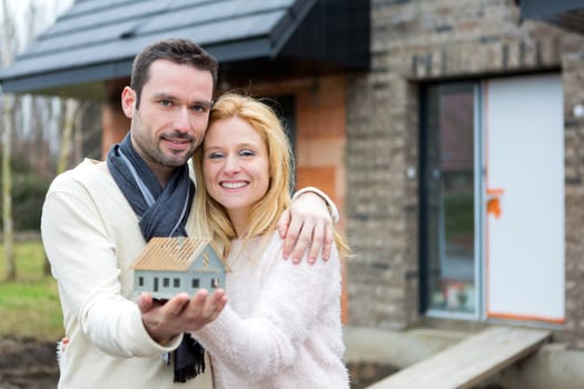 VIew of a Young happy couple handling a miniature house in front of their future home