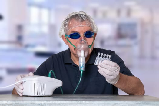 Coronavirus treatment concept. Man at table with nebulizer mask, safety goggles and medical hair hood and gloves looks at the ampoules over blurred hospital background. Protection of the respiratory tract.