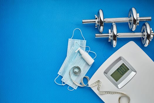 Sport equipment, scales, medical masks and sanitizer on blue background. Top view, copy space. Fitness.  Isplation period. Weight and health control during quarantine. Concept of covid or coronavirus
