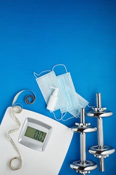 Sport equipment, scales, medical masks and sanitizer on blue background. Top view, copy space. Fitness.  Isplation period. Weight and health control during quarantine. Concept of covid or coronavirus
