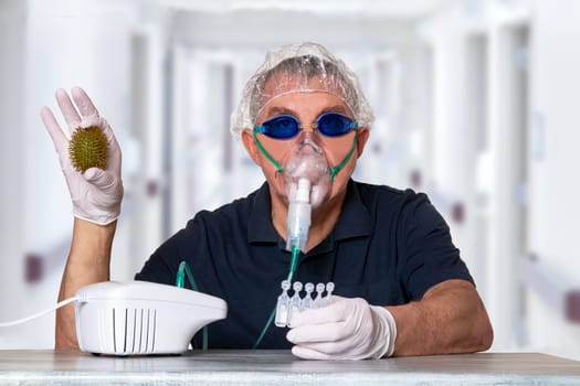 Coronavirus treatment concept. Man at table with nebulizer mask, safety goggles and medical hair hood and gloves shows an abstract model of a virus covid-19 and ampoules over blurred hospital background.