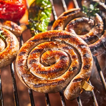 Coil of spicy beef sausage grilling on a BBQ over the coals with fresh tomatoes and chili peppers in a close up square view for advertising