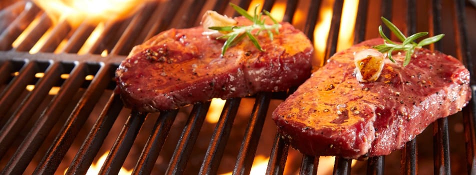 Panorama banner with raw beef steak on a BBQ grill sizzling over flaming hot coals in a close up view with copy space