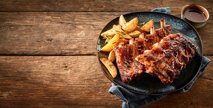 Barbecued ribs and wedges with a side dish of sauce on a rustic timber table top.