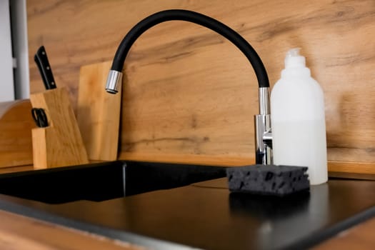 Side view of a black granite kitchen sink with a flexible faucet on which there is dish detergent and a black sponge against the background of a wooden countertop on which kitchen utensils are placed. Kitchen island, dishwashing, kitchen cleanliness. High quality photo