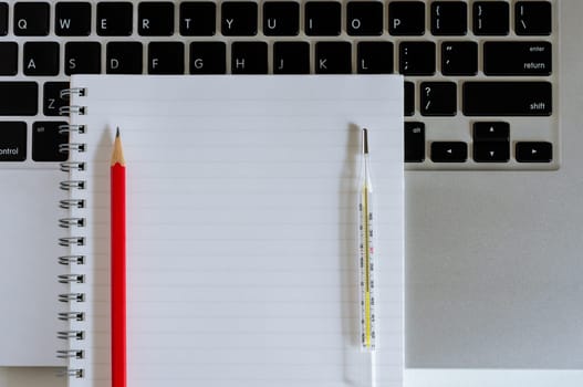 Blank notebook with red pencil on laptop and medical thermometer on laptop. Work from home to protect from coronavirus during the COVID-19 quarantine period